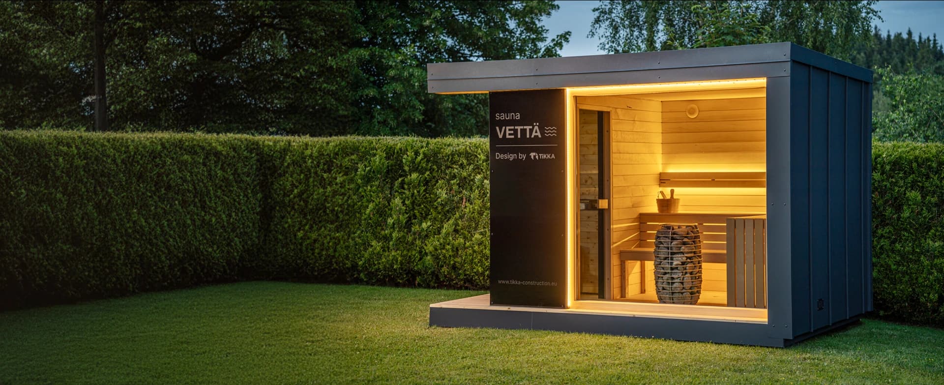 TIKKA Vettä garden sauna at dusk – lit interior with benches and heater behind the glass wall