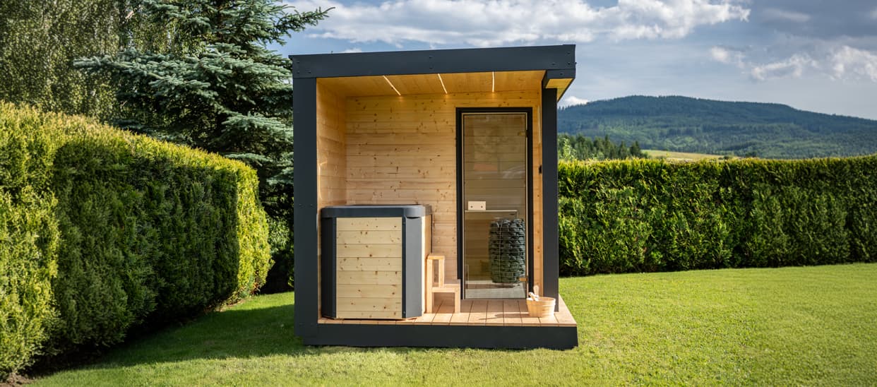 TIKKA garden sauna in a garden – anthracite cabin with a wooden deck against a hedge